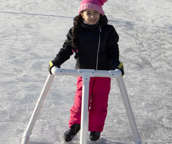 a young girl bundled up in warm clothing, wearing ice skates on an outdoor ice rink while holding onto a skate helper