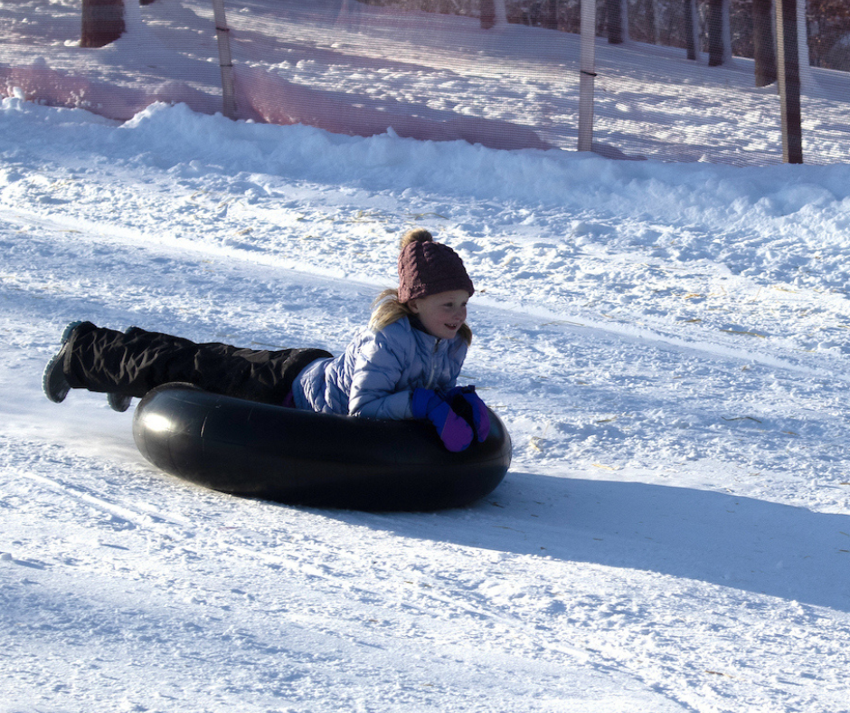 a young girl bundled up in warm clothing, laying on a snow tube and going down the sledding hill